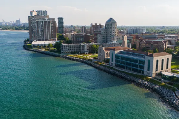 An aerial perspective of the university's lake shore campus. The afternoon sun illuminates the buildings casting long shadows across the campus and Lake Michigan's emerald water..