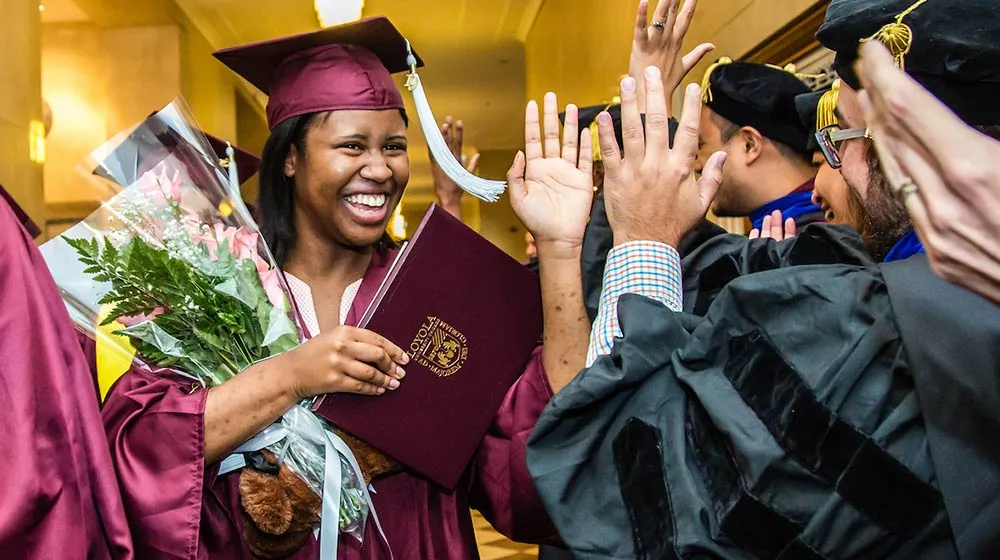 A smiling graduate in a maroon cap and gown holds a diploma and a bouquet of flowers while receiving high-fives from faculty or fellow students 