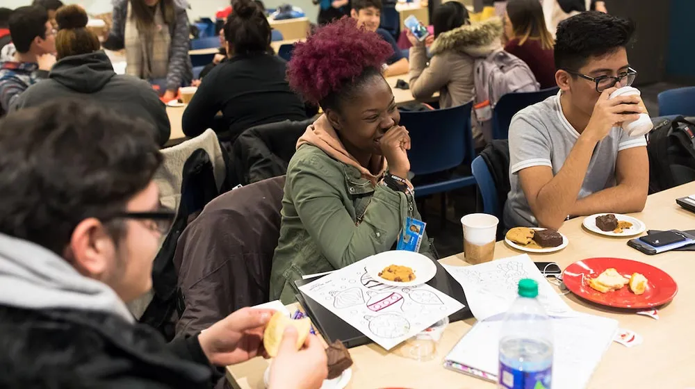 Smiling students laughing and talking together at a table with snacks and coffee at the Water Tower Campus.