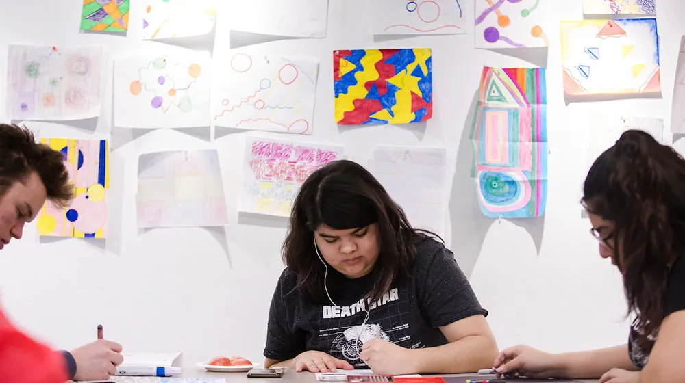 Students working on art projects at a table in front of a wall of drawings.