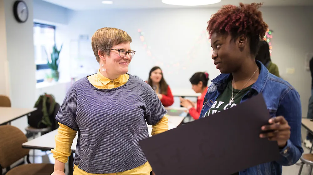 Two people smiling and talking in a classroom, one holding a large black poster board