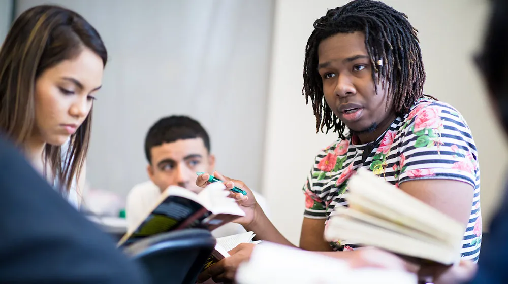 Students engaged in a small group discussion, one speaking while holding a book and pen