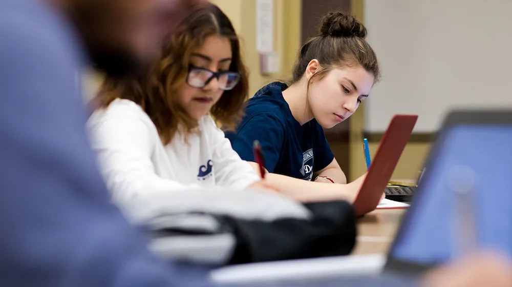Two students focused on their coursework during a class at the Water Tower Campus, with laptops and notebooks on the desk.