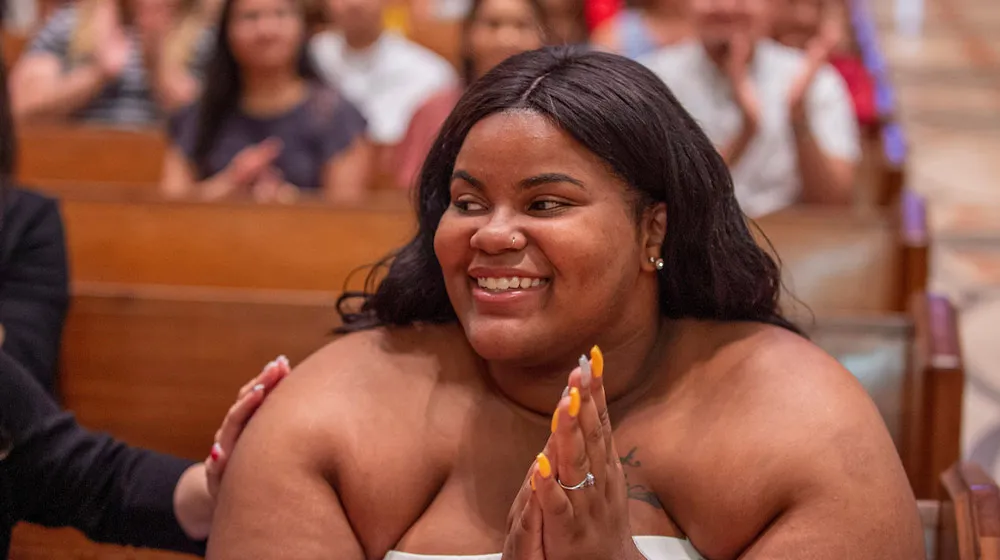 A smiling young woman sits during mass, hands pressed together in a celebratory gesture as someone places a supportive hand on her shoulder.
