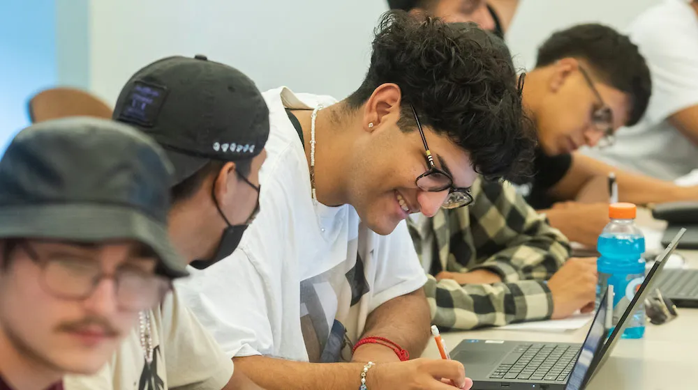A student with curly hair and glasses smiles while taking notes on a laptop, capturing a positive and productive atmosphere at Water Tower Campus.