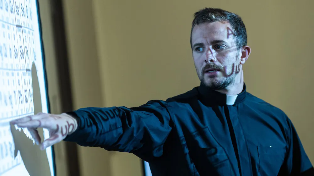 A Jesuit priest pointing to a periodic table projected onto a screen during a presentation at the Water Tower Campus.