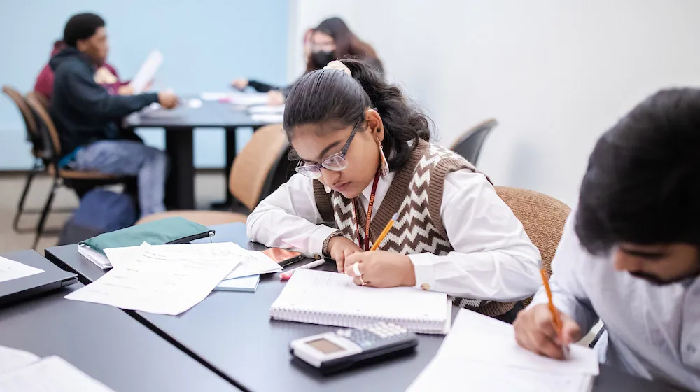 A student taking notes during a class session at the Water Tower Campus, with a calculator and papers on the desk.