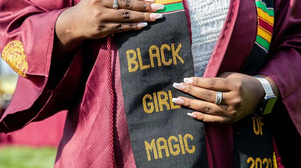A close-up of a graduate's hands resting on a Kente-style graduation stole that reads 