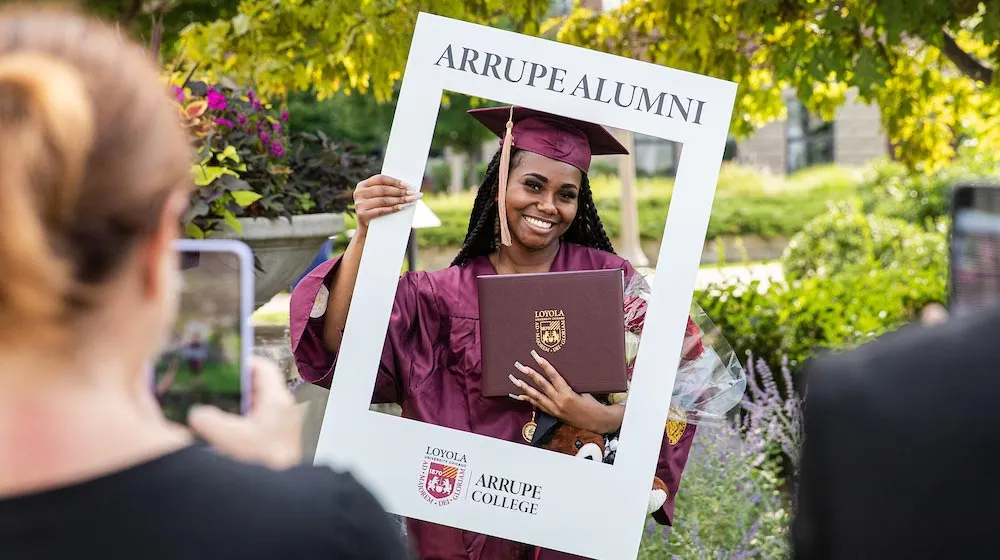 A smiling graduate in a maroon cap and gown holds a diploma while posing inside a white 