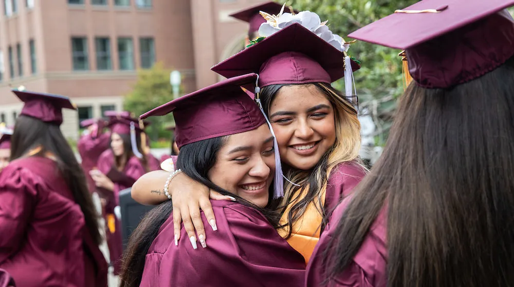 Two Arrupe graduates in maroon caps and gowns share a warm embrace and smile during a commencement ceremony outdoors.
