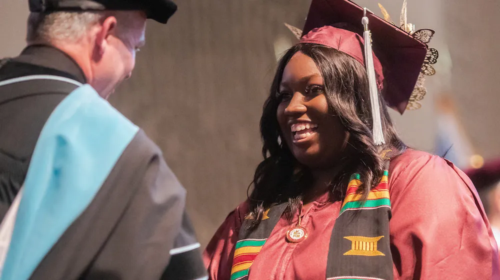 A joyful Black woman celebrating her graduation in maroon regalia. She wears a traditional Kente stole over her gown and a decorated mortarboard.