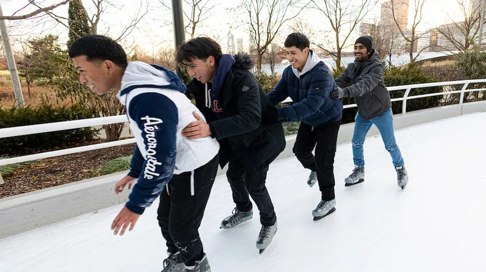 Four students ice skating together in a line at the Maggie Daley Park Skating Ribbon, with the Chicago skyline visible in the background.
