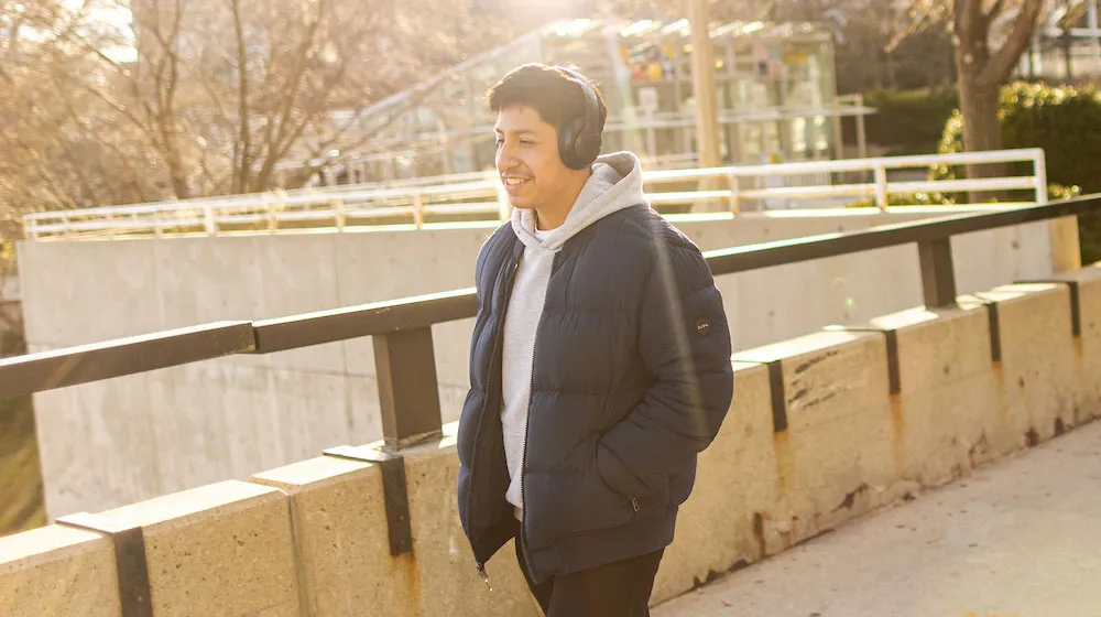 Smiling young man walking outside with headphones and a puffer jacket.