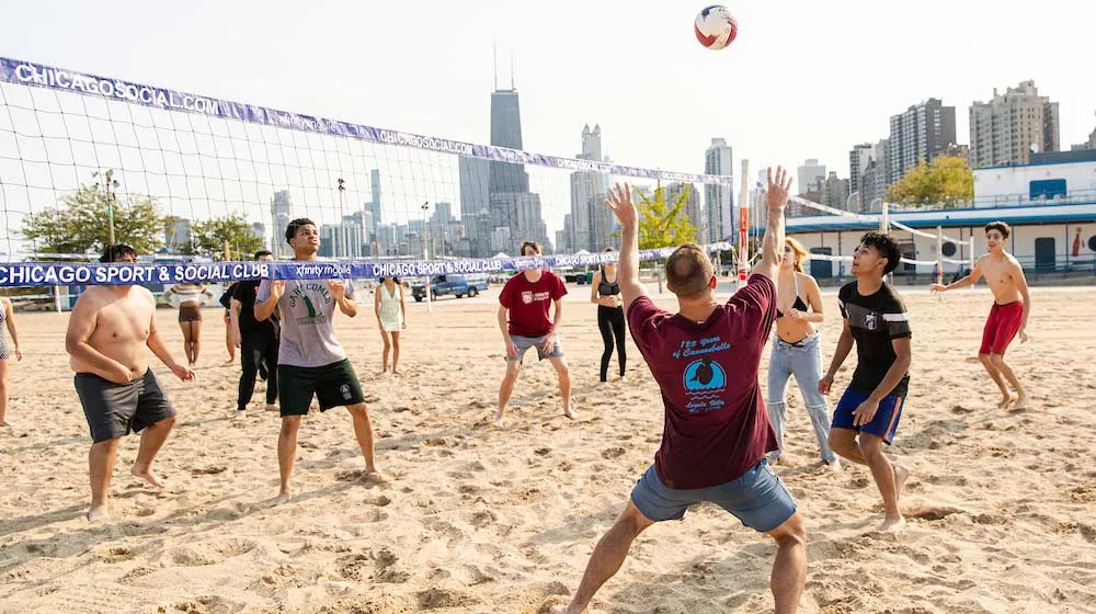 Students playing beach volleyball at North Avenue Beach, with the iconic 875 North Michigan Avenue building and Chicago skyline in the background.