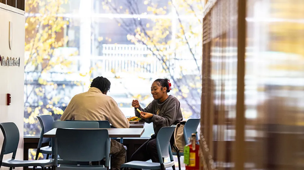 Two students eating and talking at a table in a student lounge with large windows overlooking yellow autumn leaves.