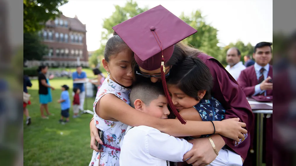 A graduate in a maroon cap and gown leans down to share a group hug with three young children on a grassy campus lawn after a commencement ceremony.