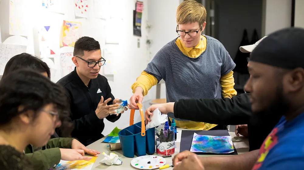 An instructor and students collaborating on an art project with various supplies on a table and colorful drawings on the wall.