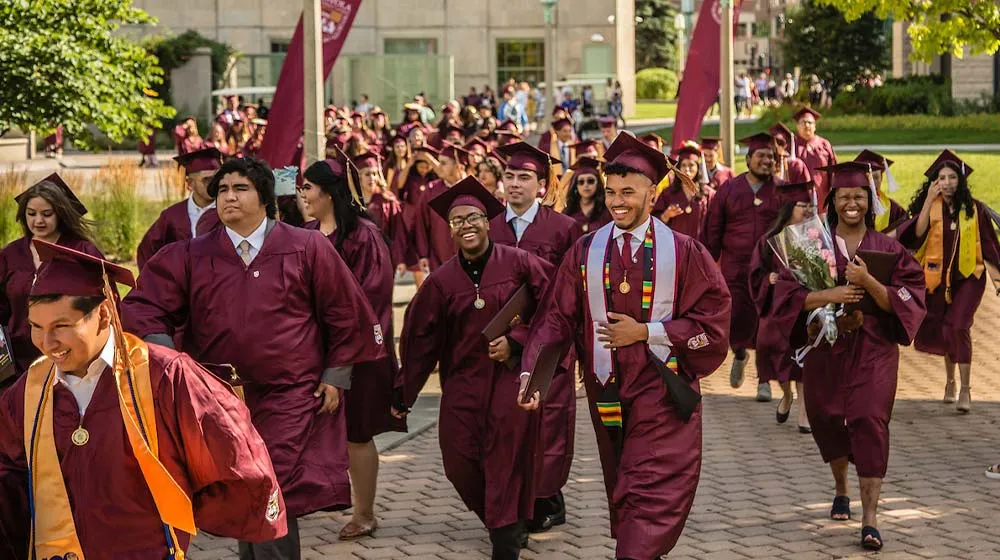 Diverse graduates in maroon regalia walk a sunny brick path on campus, smiling while wearing decorative stoles and carrying their diplomas.