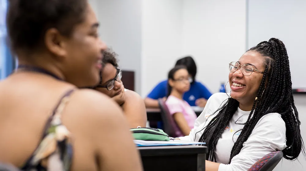 Smiling student with long braids and glasses laughing with classmates in a room.