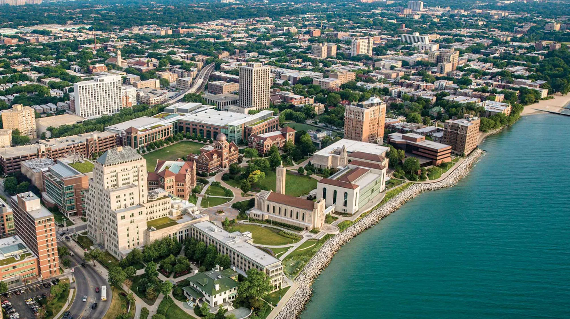 Aerial view of the Lakeshore campus with large academic buildings, green lawns, and surrounding city neighborhoods extending into the distance.
