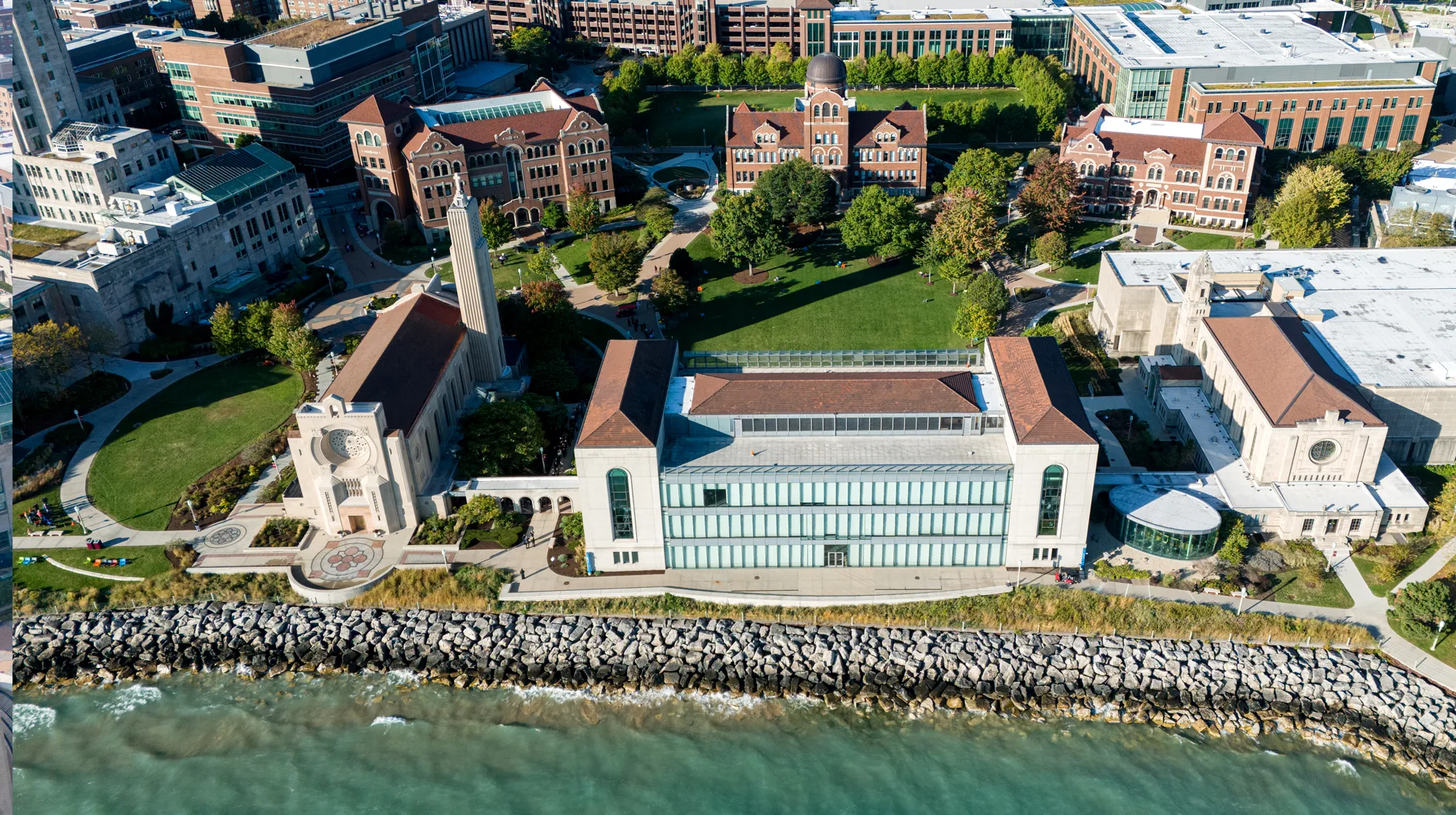 Aerial view of a lakeside campus featuring a large glass‑front building, surrounding historic structures, green lawns, and walkways near the shoreline.