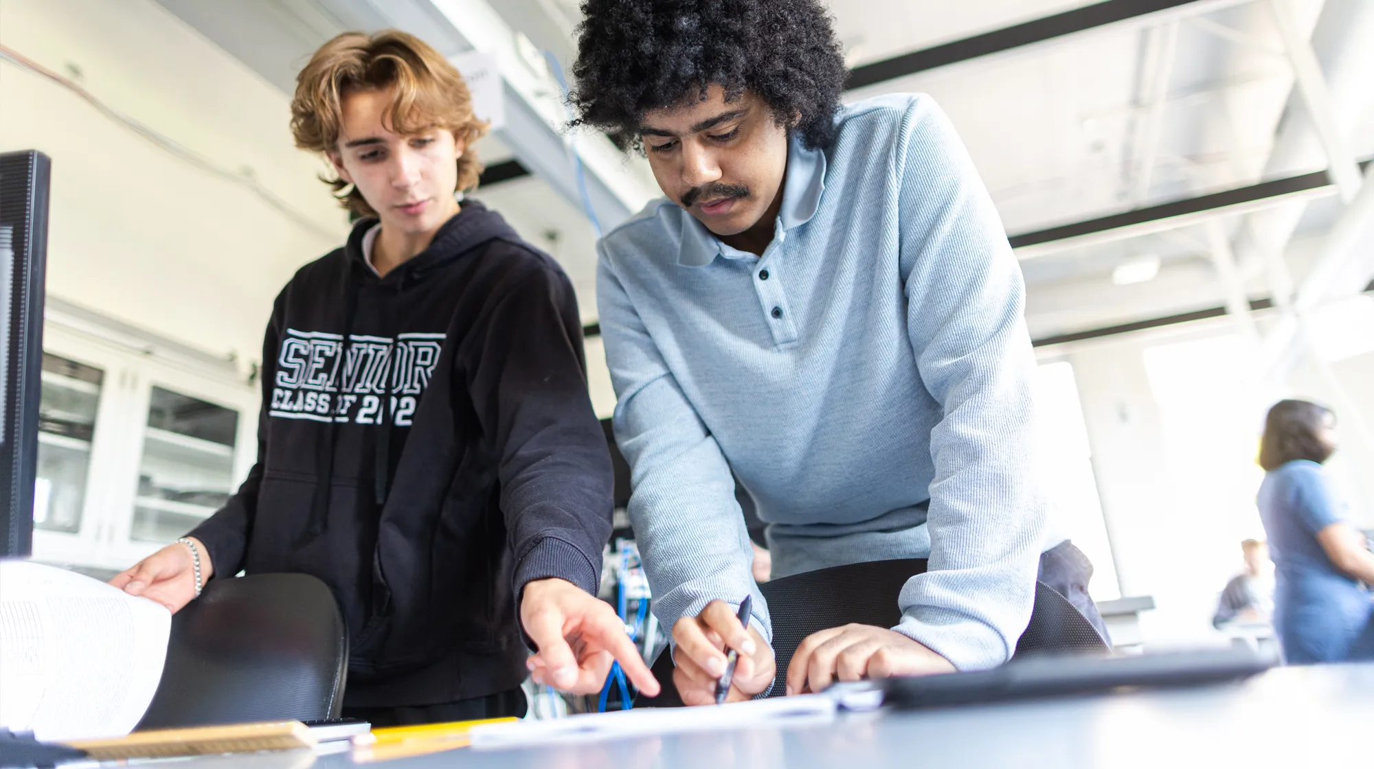 Two students work together at a desk in a bright classroom, focusing on a shared task while standing beside computer equipment.