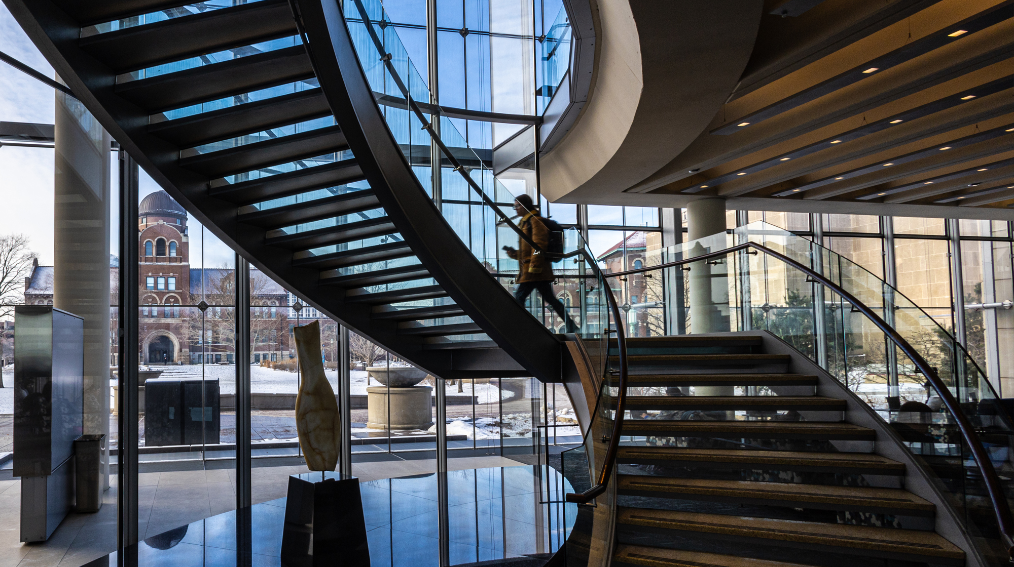 A person walks up a curved glass staircase inside a bright atrium, with large windows revealing snowy campus buildings and trees outside.
