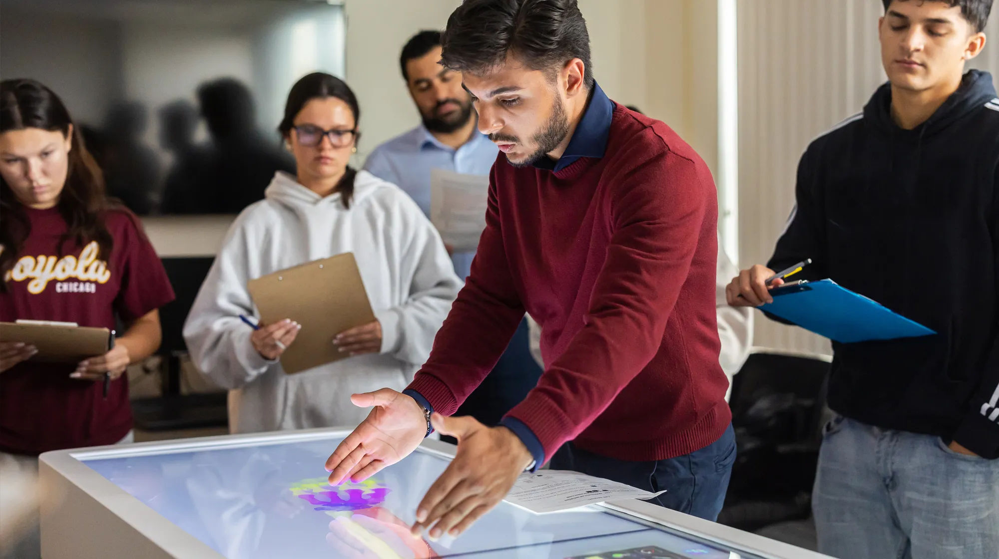 Instructor demonstrating an interactive touchscreen table while a group of students observe and take notes on clipboards in a classroom setting.
