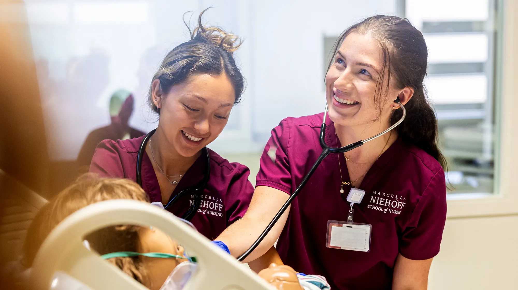 Nursing students in matching scrubs work together beside a hospital bed, using a stethoscope and reference sheets during a clinical training simulation.