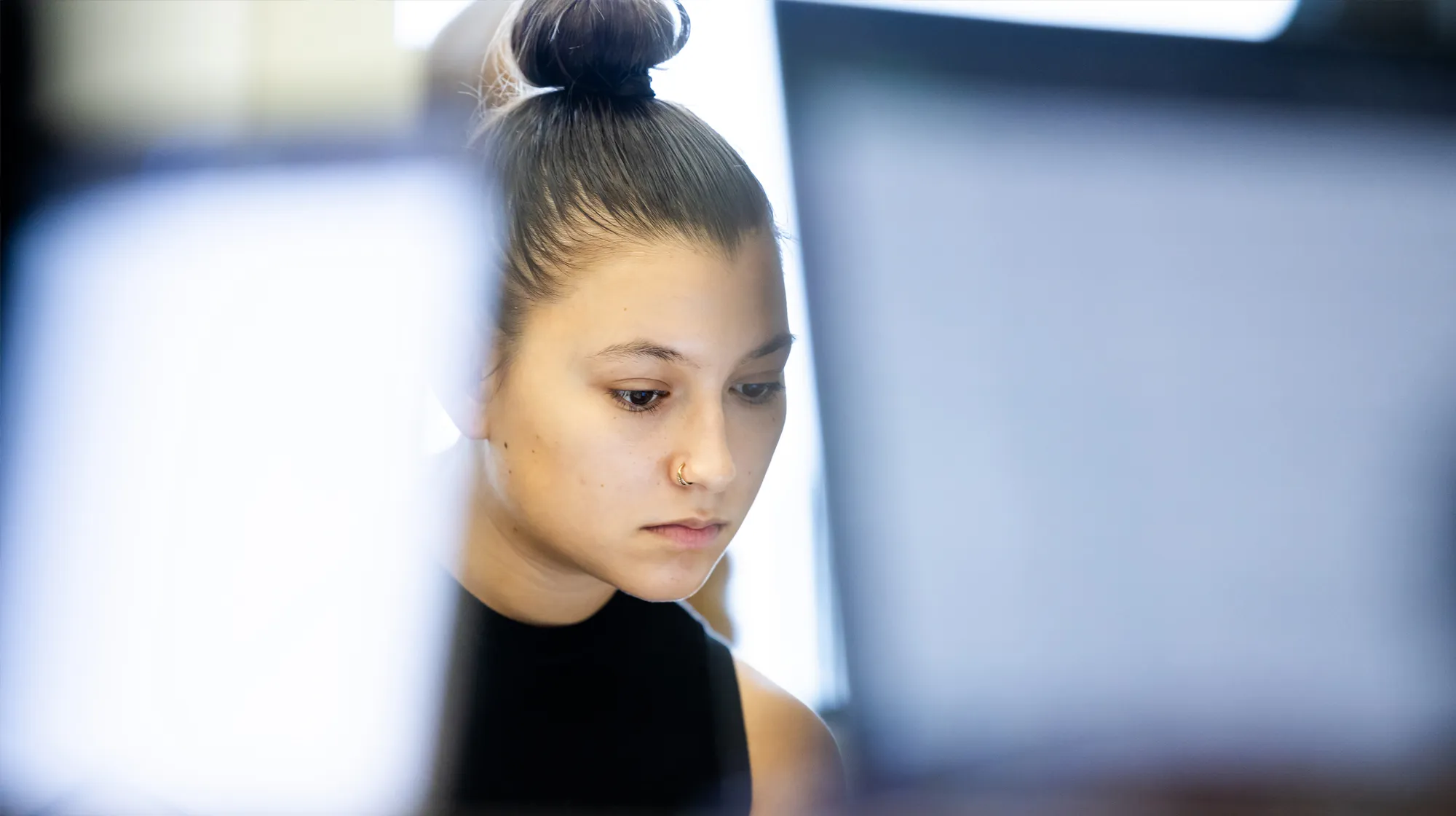 A person sits behind two bright laptop screens in a softly lit space, with hair in a high bun and background light visible.