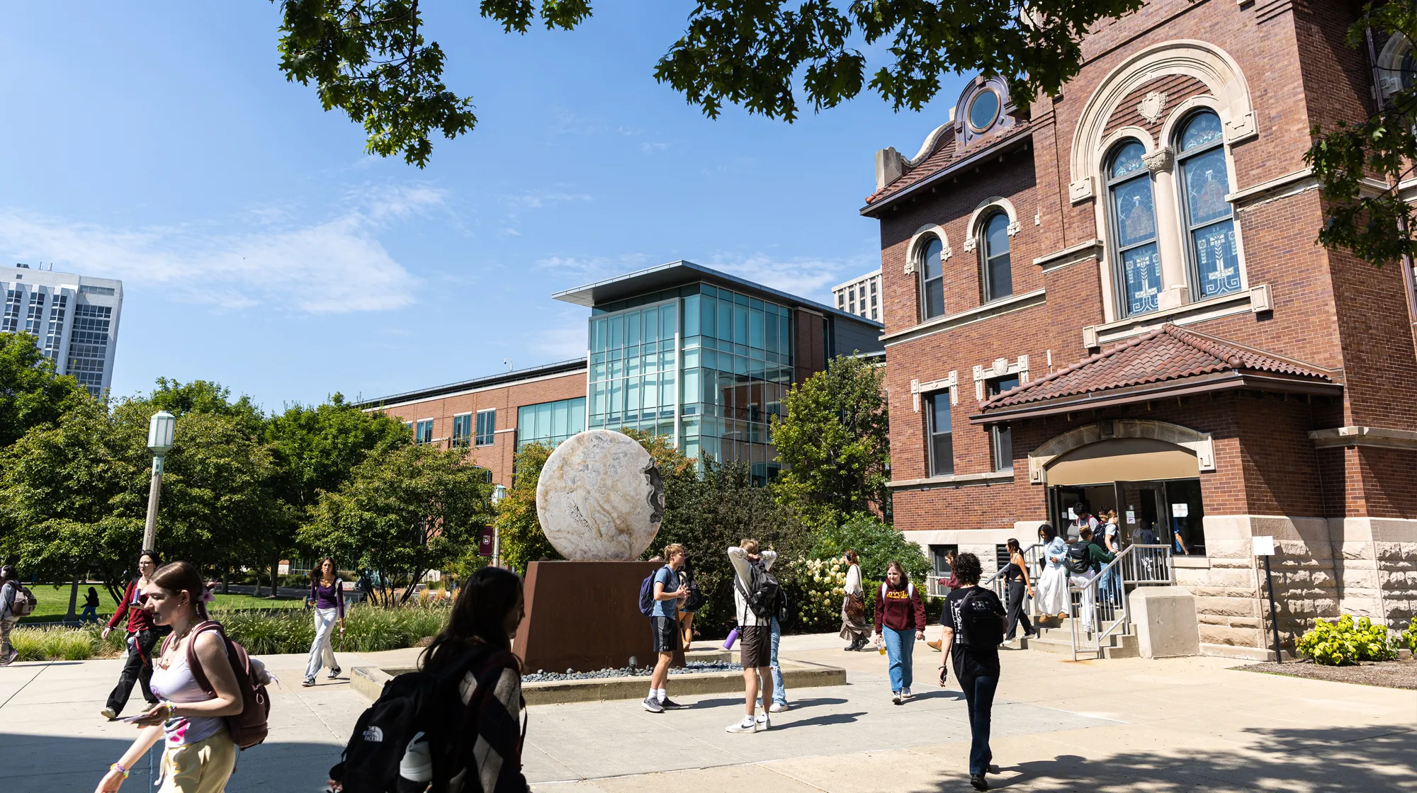 Students walk across a sunny campus plaza near a large spherical sculpture, with modern and historic academic buildings surrounded by trees in view.