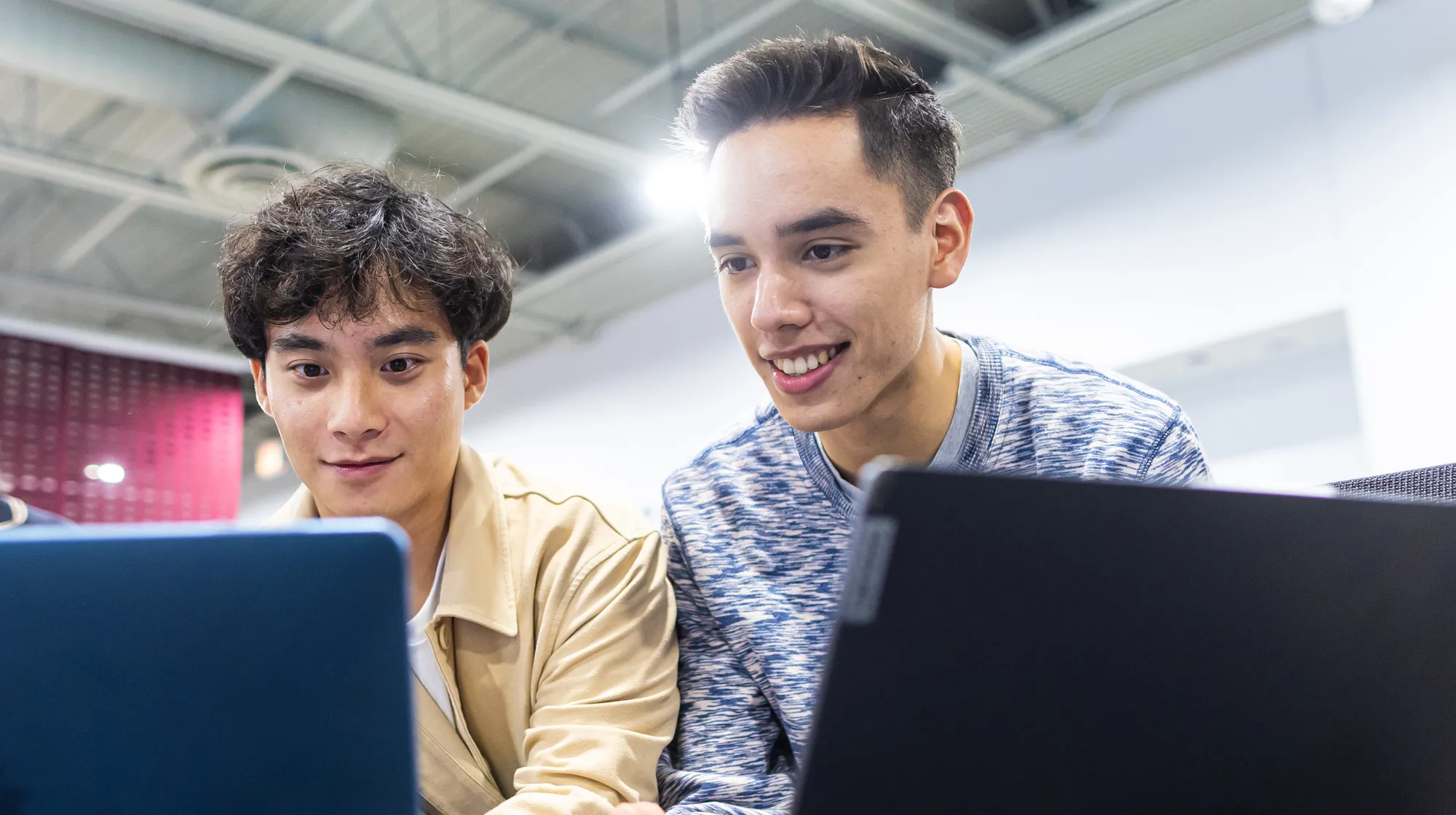 Two students sit closely together working on laptops in a bright indoor space, focusing on their screens beneath overhead industrial-style lighting.