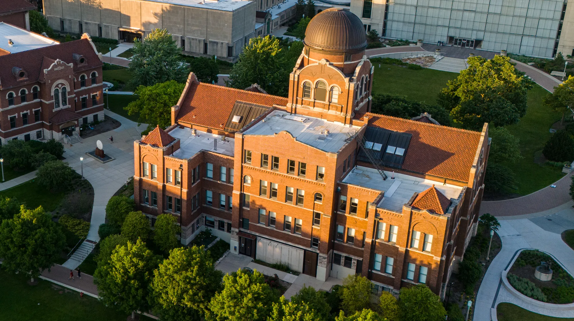 Aerial view of a historic red‑brick campus building with a prominent domed tower, surrounded by trees, pathways, and nearby academic structures.