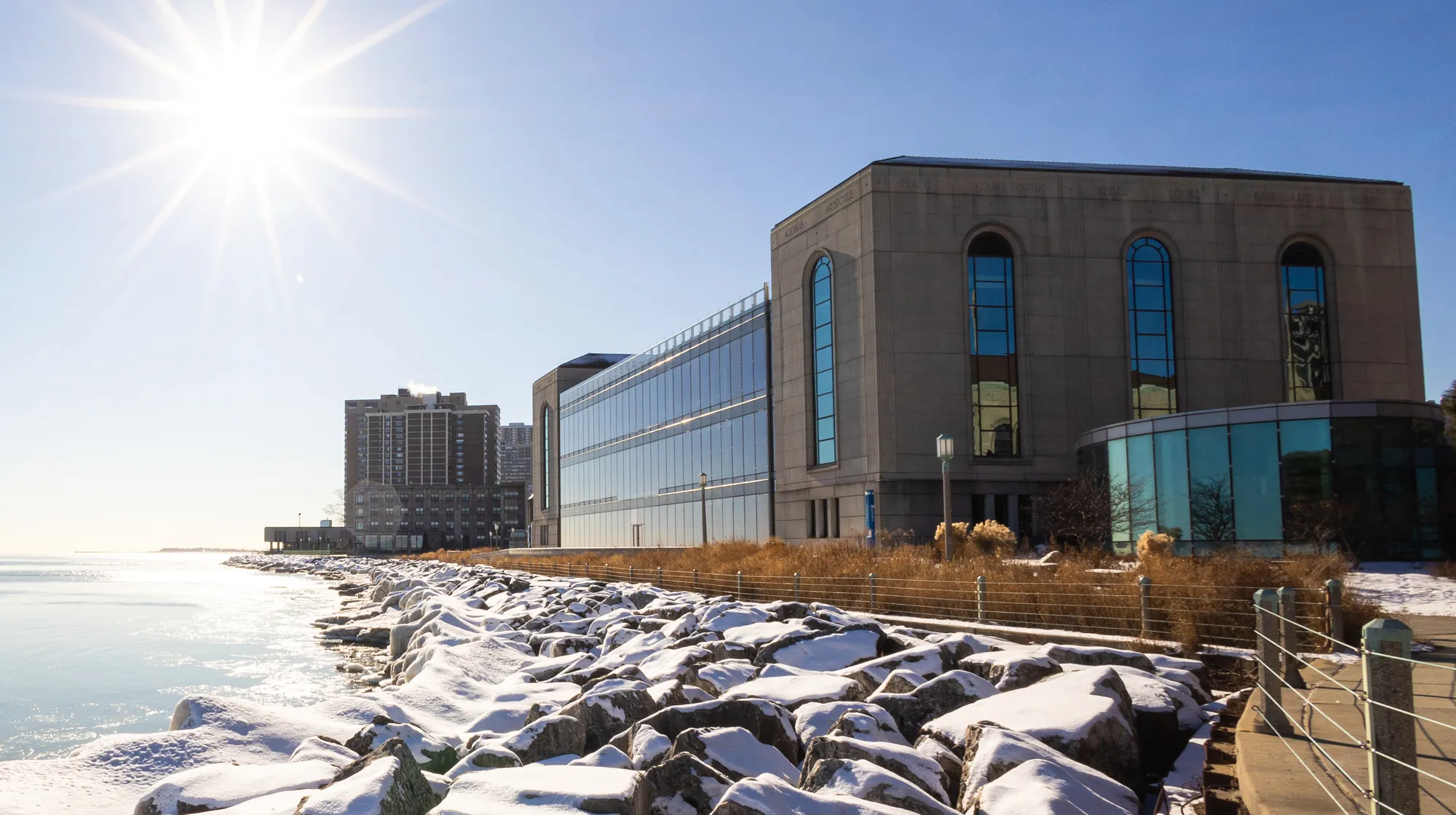Sunlit winter view of a modern lakeside building beside snow‑covered rocks, with calm water, blue sky, and nearby high‑rise structures.