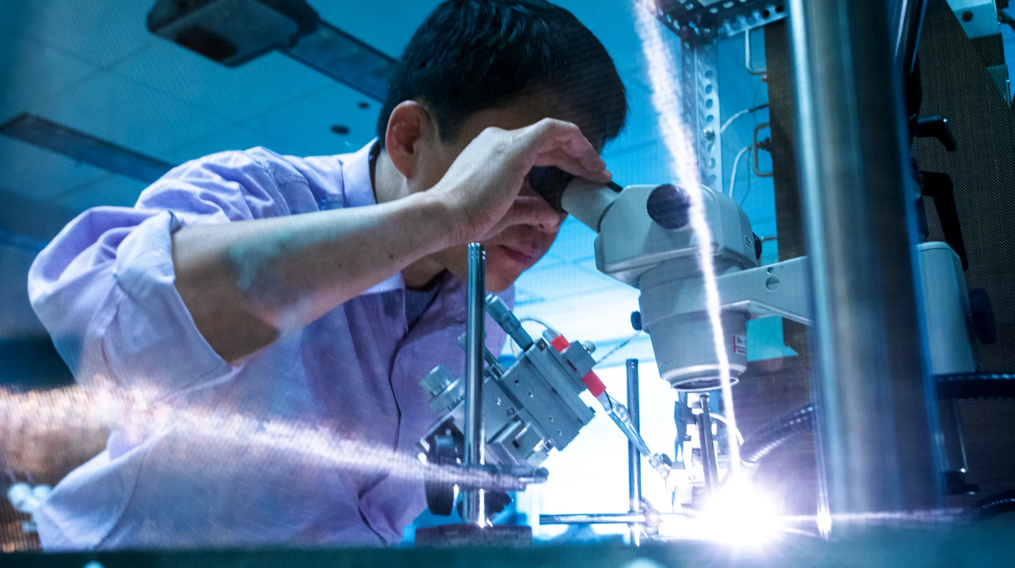 Researcher operating laboratory equipment while examining a sample through a microscope.