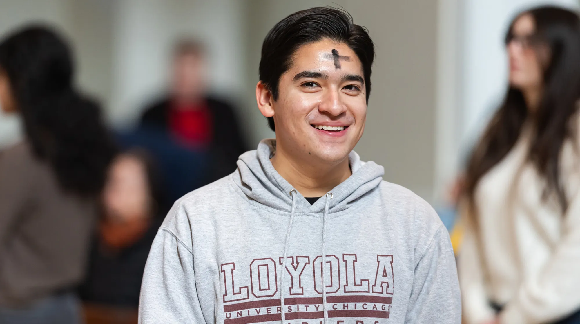 Student wearing Loyola hoodie with ash cross on forehead, smiling indoors