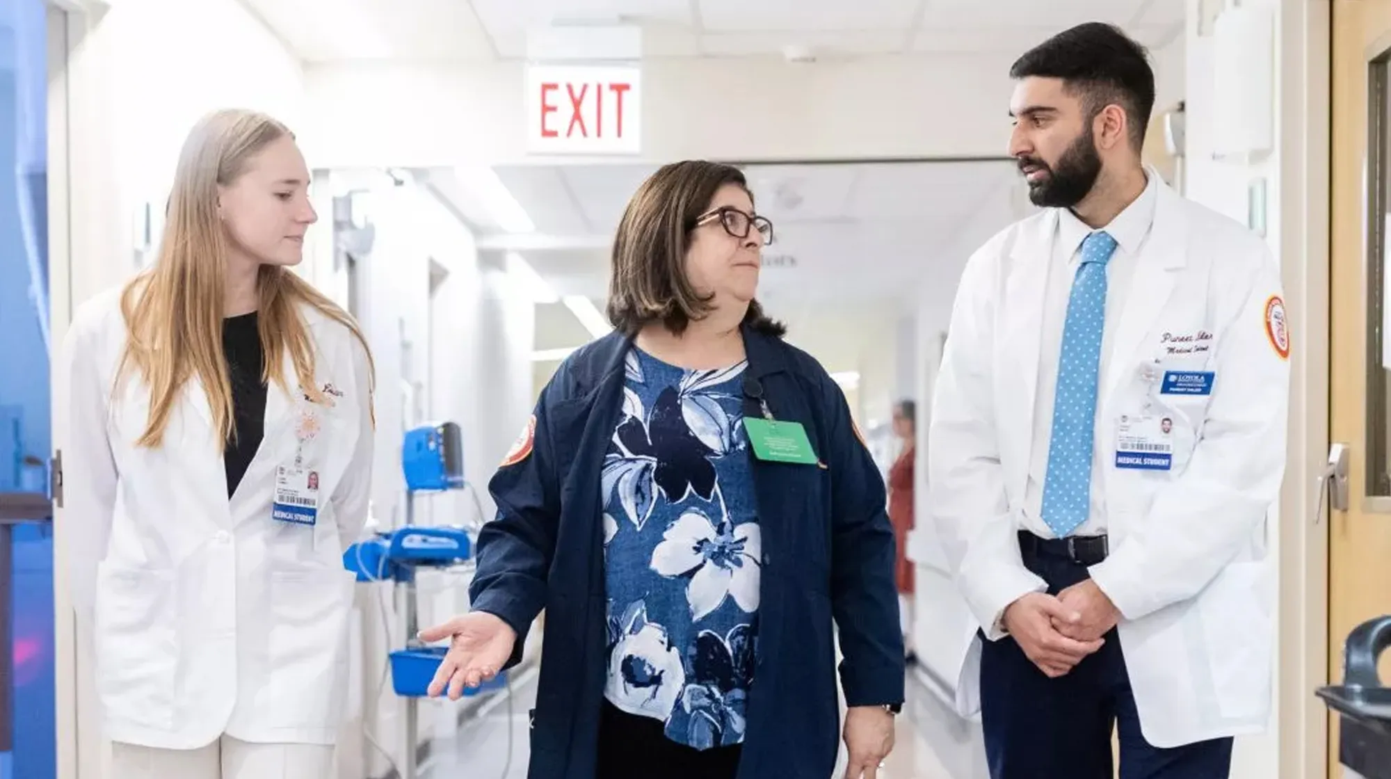 Two medical students in white coats walk alongside a staff member, talking in a hospital corridor
