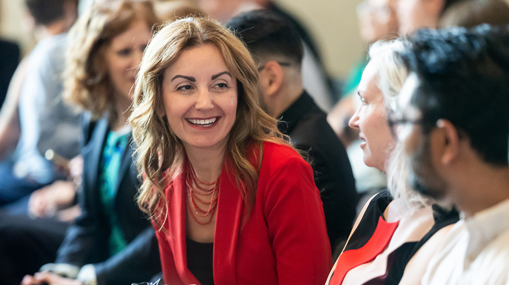 A smiling woman with a bright red jacket interacts with two people, a man and a woman, sitting on her left A smiling woman with a bright red jacket interacts with two people, a man and a woman, sitting on her left