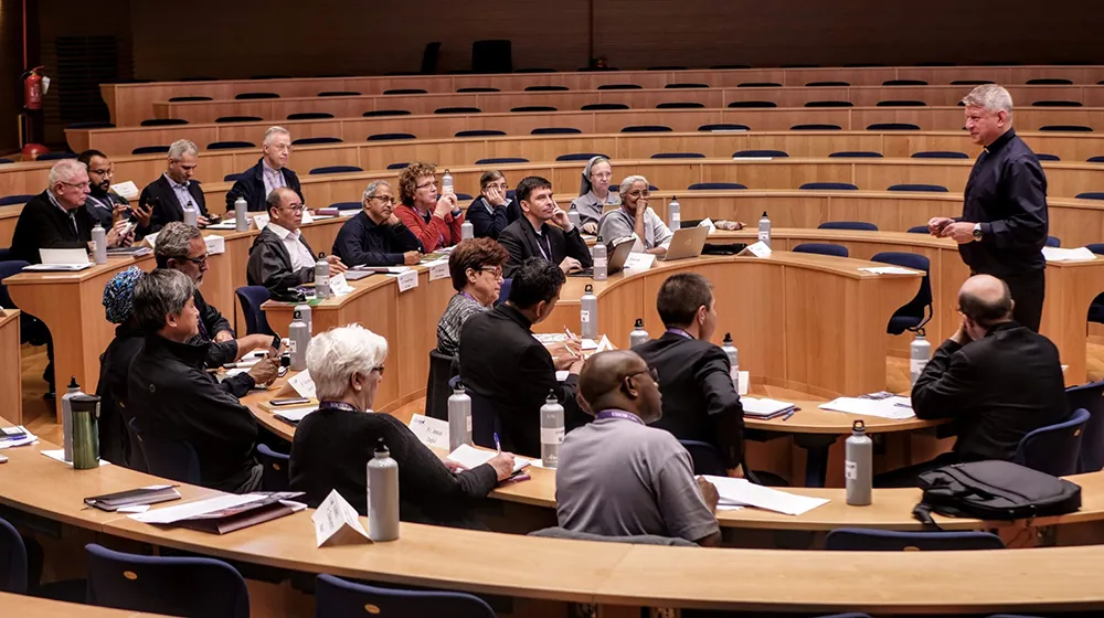 A variety of several people sit in a circular sitting arrangement listening to a presenter at a podium. A variety of several people sit in a circular sitting arrangement listening to a presenter at a podium.