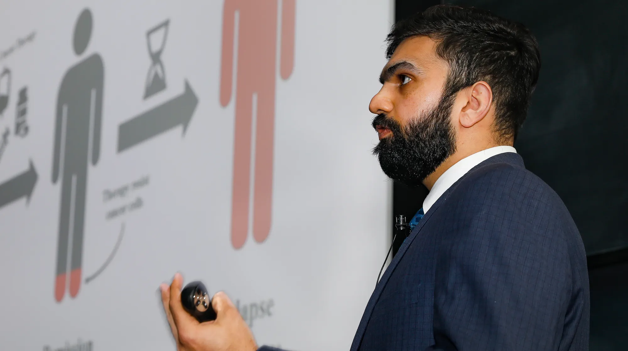 A man in a suit presents in front of a slide with icons and arrows, gesturing while speaking