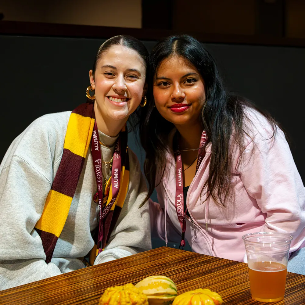 Two alumnae wearing maroon and gold striped scarves chat at an Alumni Weekend event.
