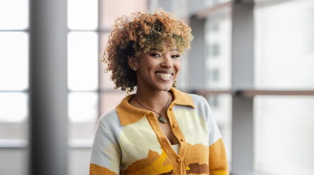 Arrupe College student with short curly hair standing indoors near large windows, wearing a patterned yellow and tan cardigan and looking confidently 