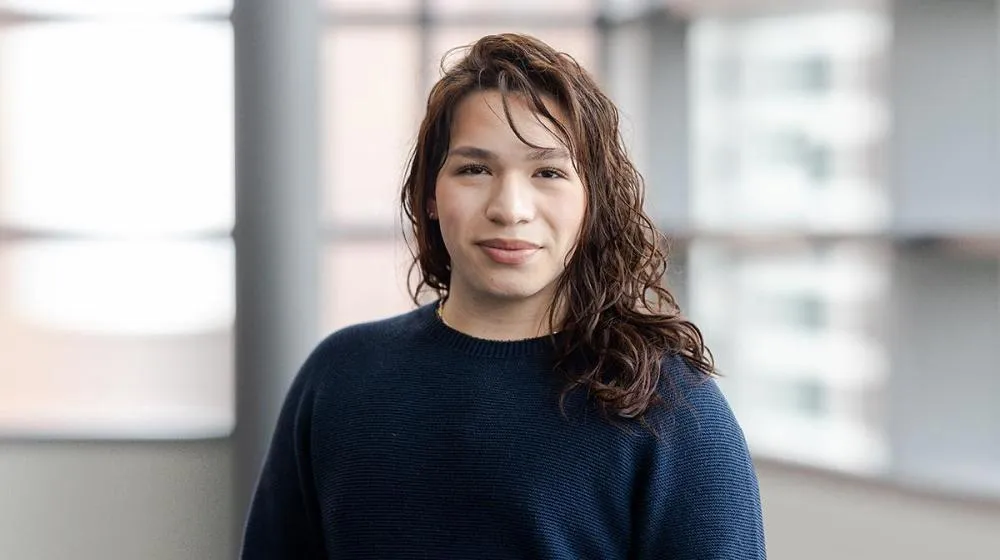 Arrupe College student standing indoors in front of large windows, wearing a navy sweater and looking confidently at the camera.