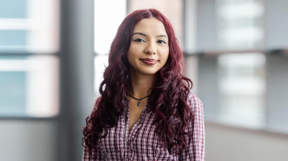 Arrupe College student with long burgundy hair standing indoors near large windows, wearing a plaid button-down shirt and looking calmly at the camera
