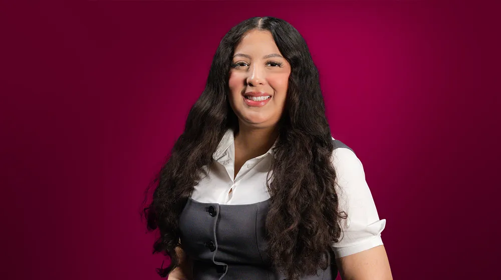 Smiling woman with long dark hair wearing a white shirt and gray vest on a maroon background