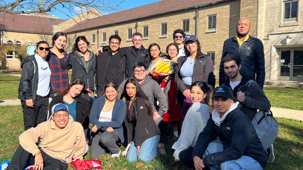 a large group of students pose, smiling outside in two rows on a sunny day 