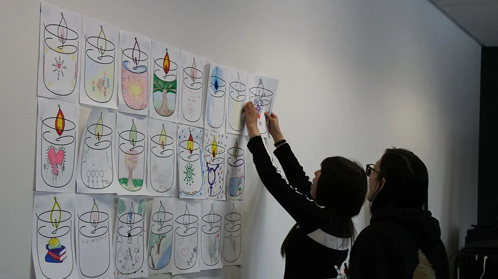 Two people adding a coloring page of a candle to a wall with other pages
