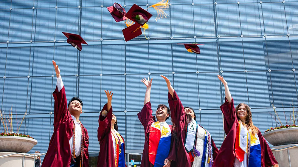 A group of 2024 graduates throw their caps in the air in celebration standing in front of the Information Commons building.