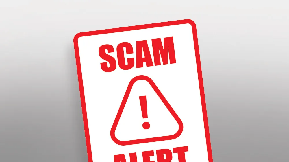 A white sign with red block letters that read SCAM ALERT. A triangle with an exclamation sign in the middle is positioned in the middle of the sign.