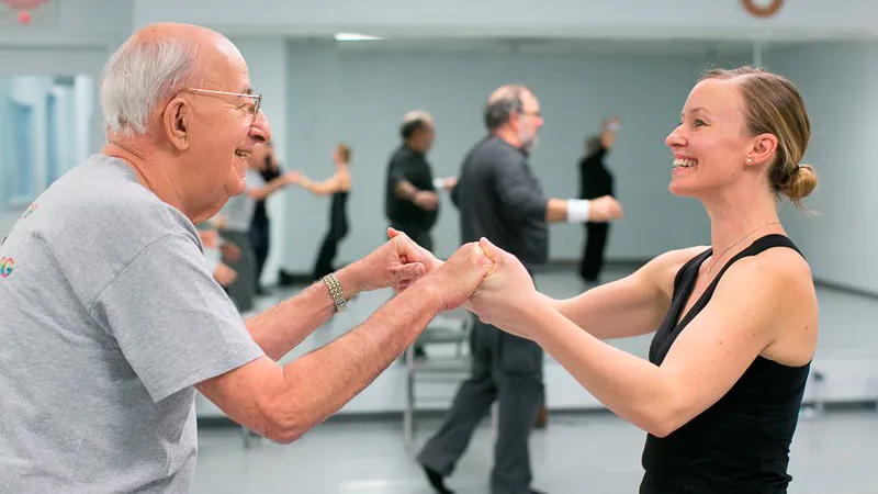 Loyola Dance Teaching Professor Sarah Cullen Fuller leading an older man through choreography in a dance studio Loyola Dance Teaching Professor Sarah Cullen Fuller leading an older man through choreography in a dance studio